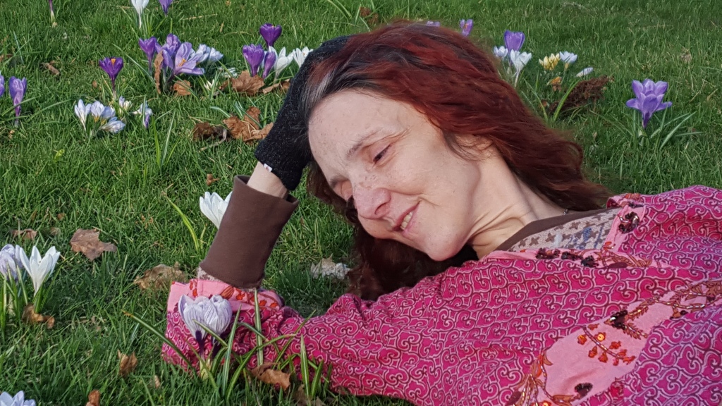 Diana Cleveland, co-leader of the EDS and CTD New England/MA Group relaxes on a lawn of fresh, green grass, surrounded by crocus flowers. She is a caucasian woman with long, red hair, and laying on her back with her head resting on her hand.