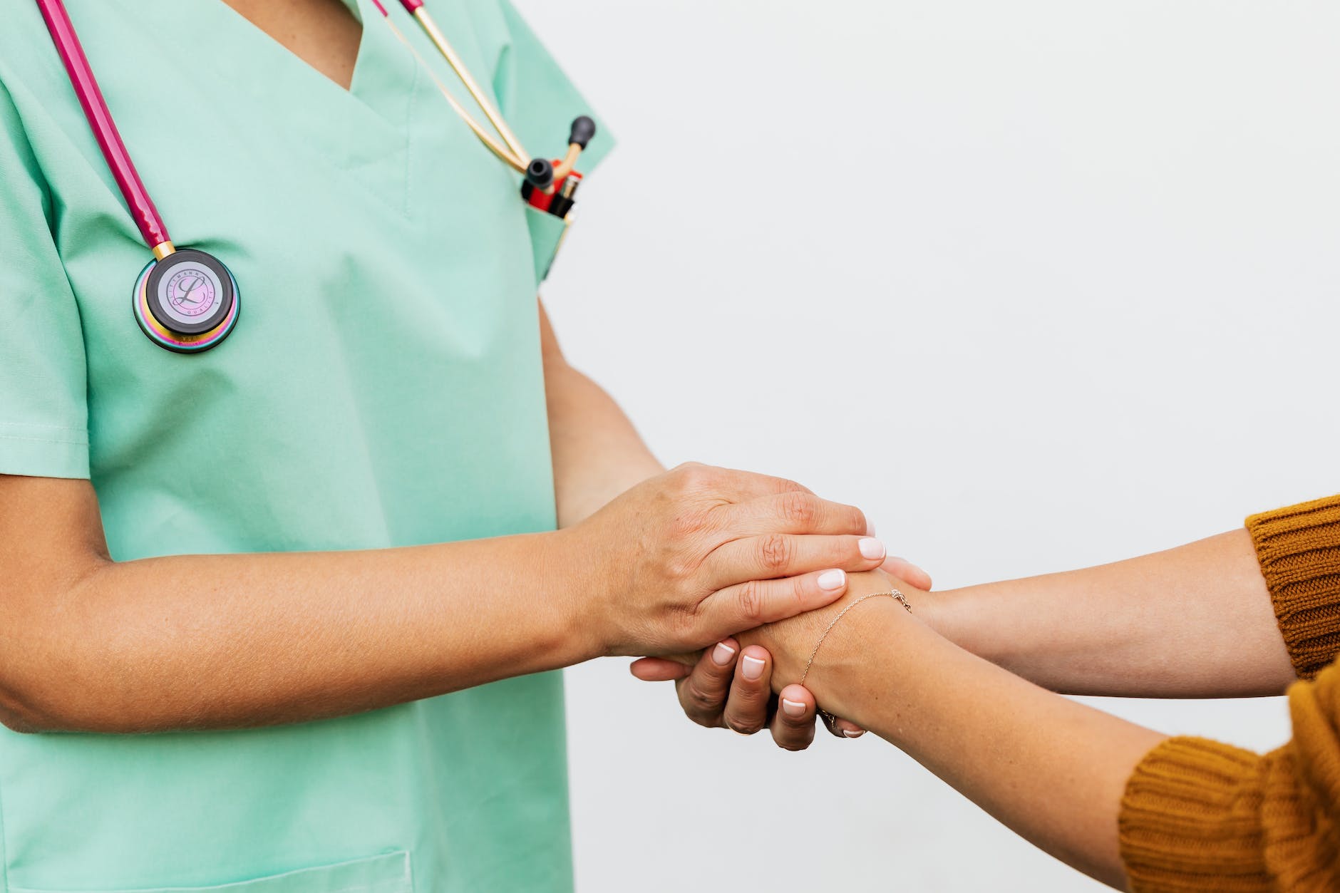 A female medical professional with a stethoscope around her neck and dressed in sage-green scrubs gently holds the hands of a woman wearing a gold-colored sweater and thin gold bracelet.