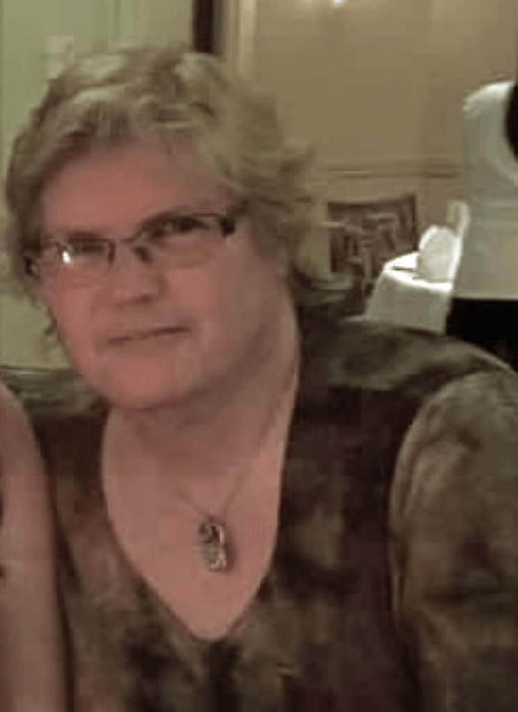 Brenda Shisslak, co-leader of the EDS and CTD New England/MA Support Group sits relaxed in a dining hall. A caucasian woman with glasses and short, wavy blonde hair, she wears a beautiful gemstone necklace.
