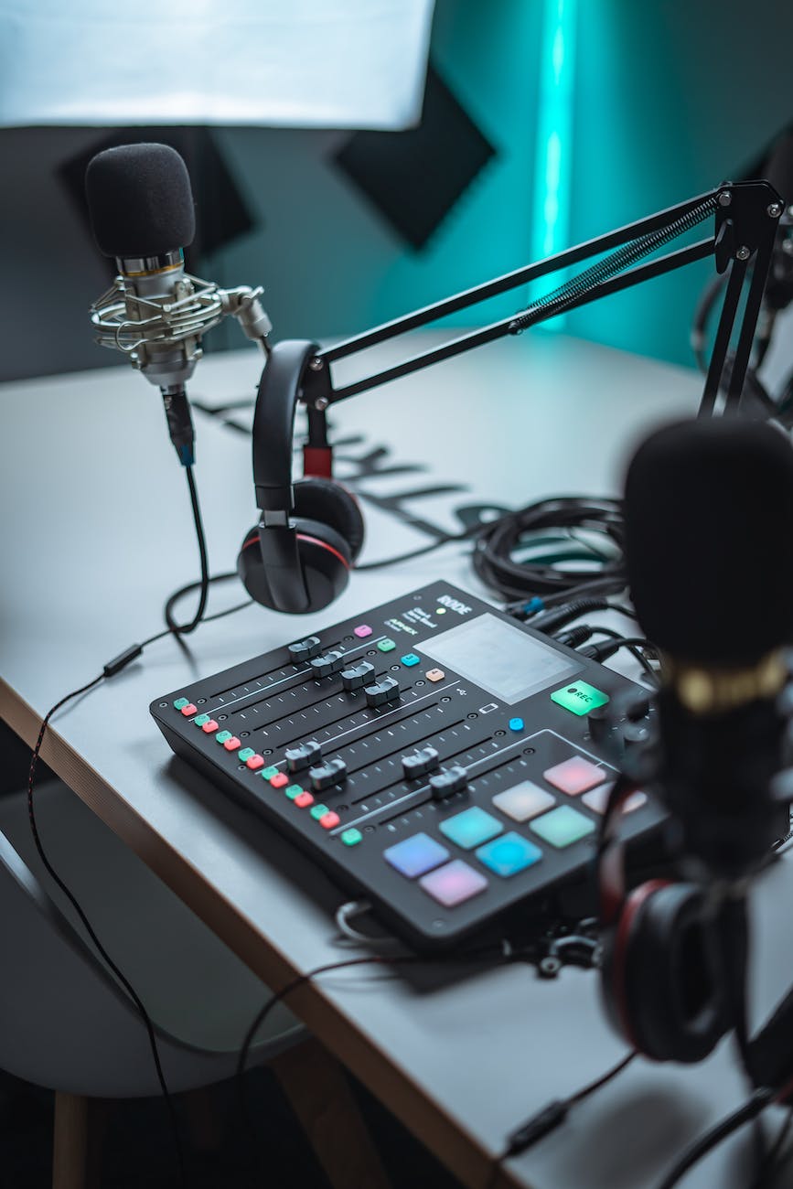 All the tools of a good podcast are set on a white table, under the glow of a blue-green neon light: microphones on a stand, equalizer, headphones, and cords.