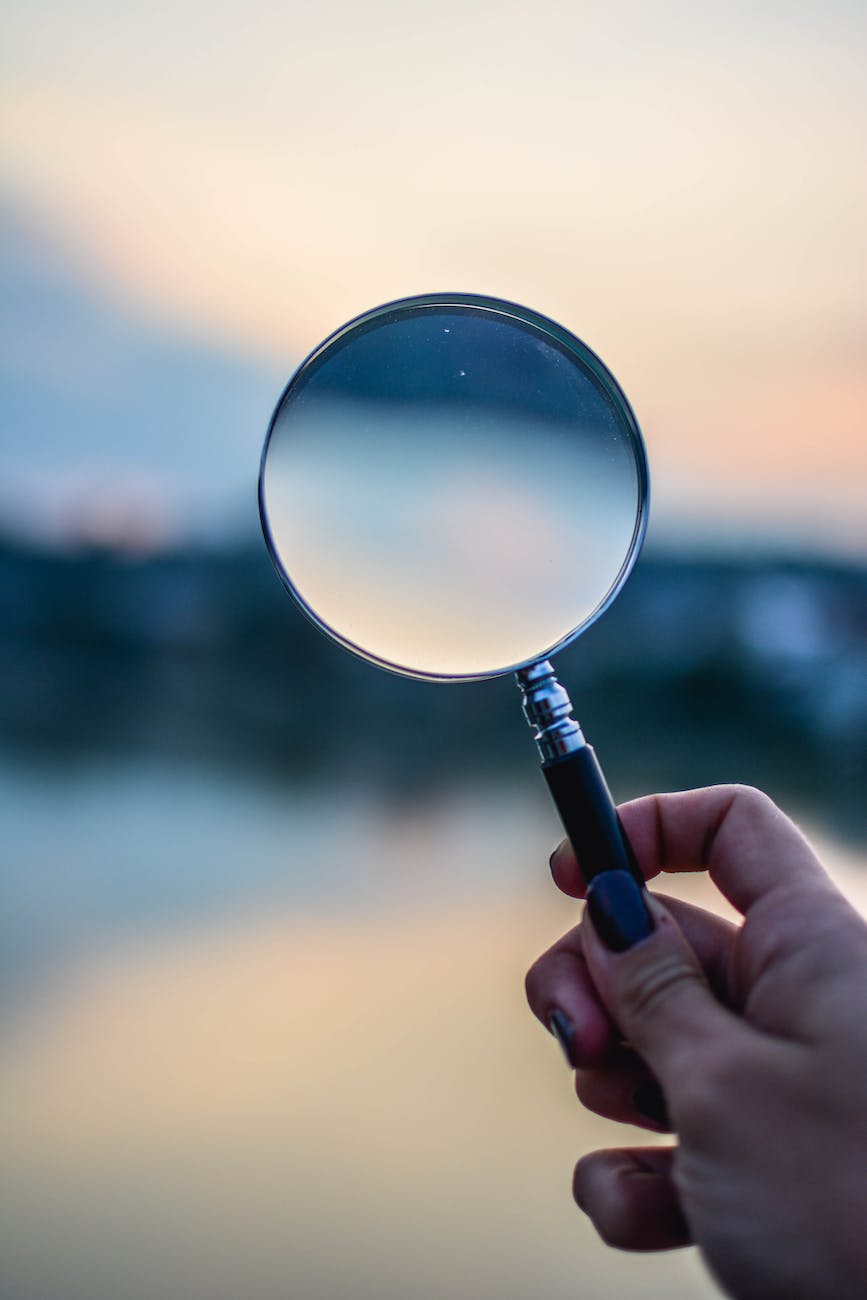 A hand with black nailpolish holds a magnifying glass before the camera. The sunset-lit background is blurry. And although the image in the glass is blurrier, it appears to reduce the image to simple values, flipping the landscape upside-down, with the dark ground at the top and the lightest bits of the sky at the bottom.